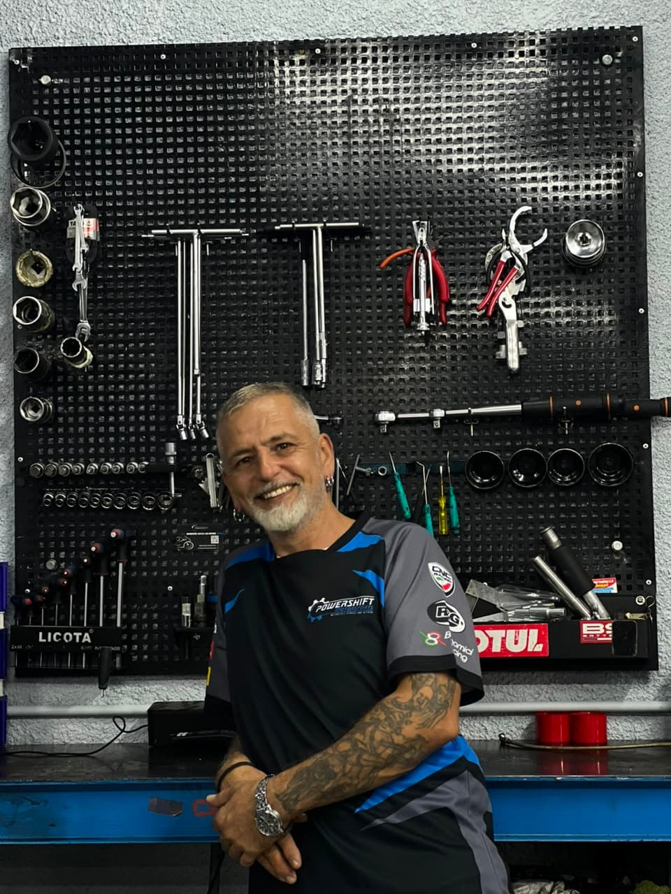 Smiling mechanic with arm tattoos stands in front of a black pegboard filled with tools.