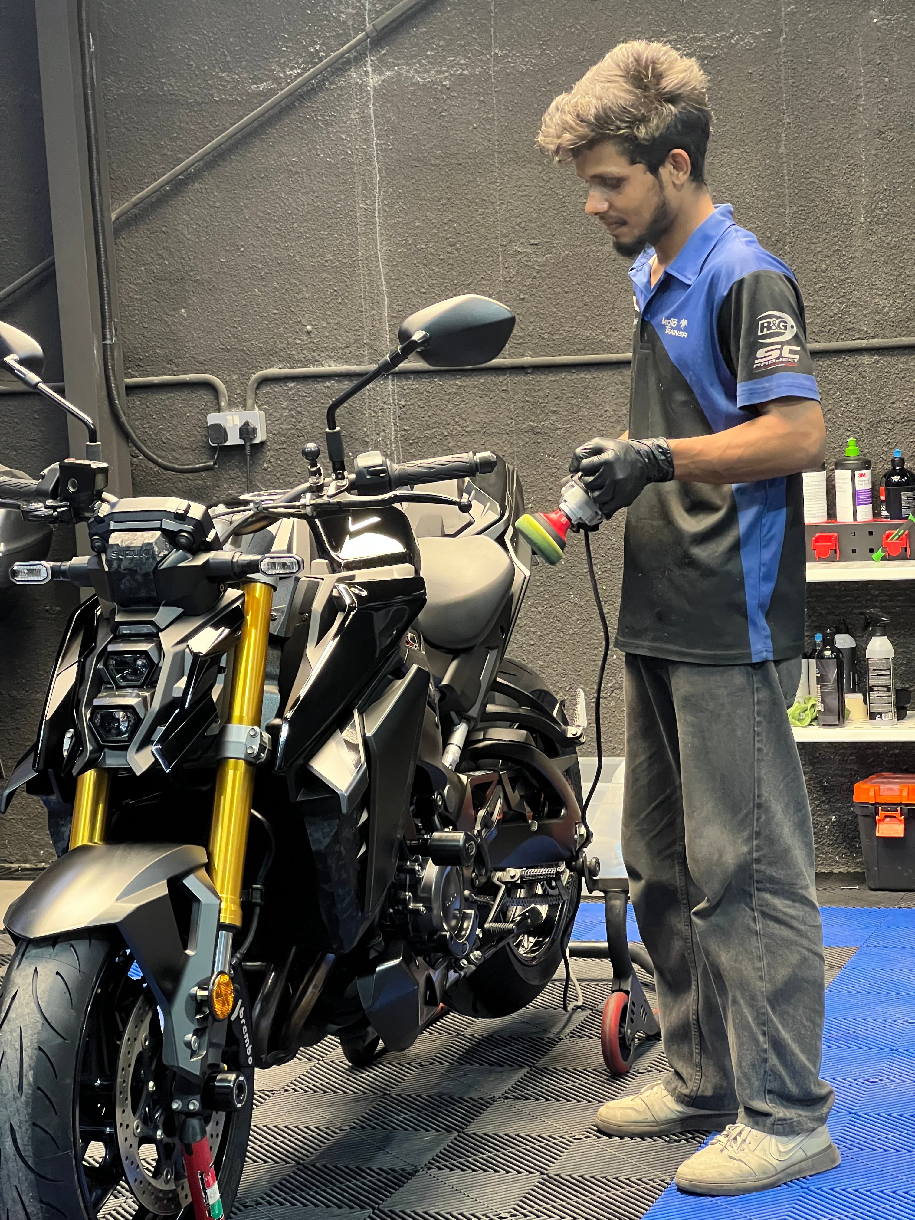 Technician using a power buffer to polish a black Suzuki motorcycle in a workshop.